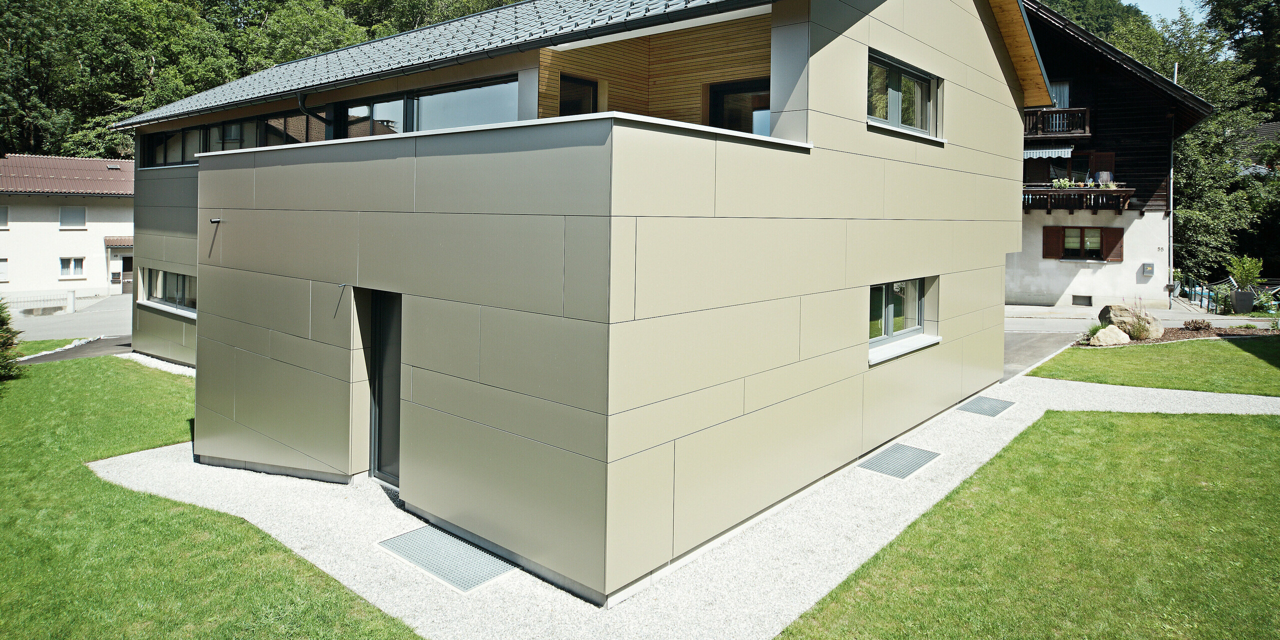 Einfamilienhaus in Hohenems mit einer stilvollen PREFA Fassade aus Aluminium Verbundplatten in Bronzemetallic und einem harmonierenden Dach aus PREFA Dachplatten in P.10 Anthrazit. Das Bild präsentiert das Gebäude in voller Pracht vor einem bewaldeten Hintergrund, wobei die saubere Linienführung und die Farbkombination der Materialien die zeitgenössische Architektur unterstreichen und für einen modernen, langlebigen und wartungsarmen Gebäudeschutz sorgen.