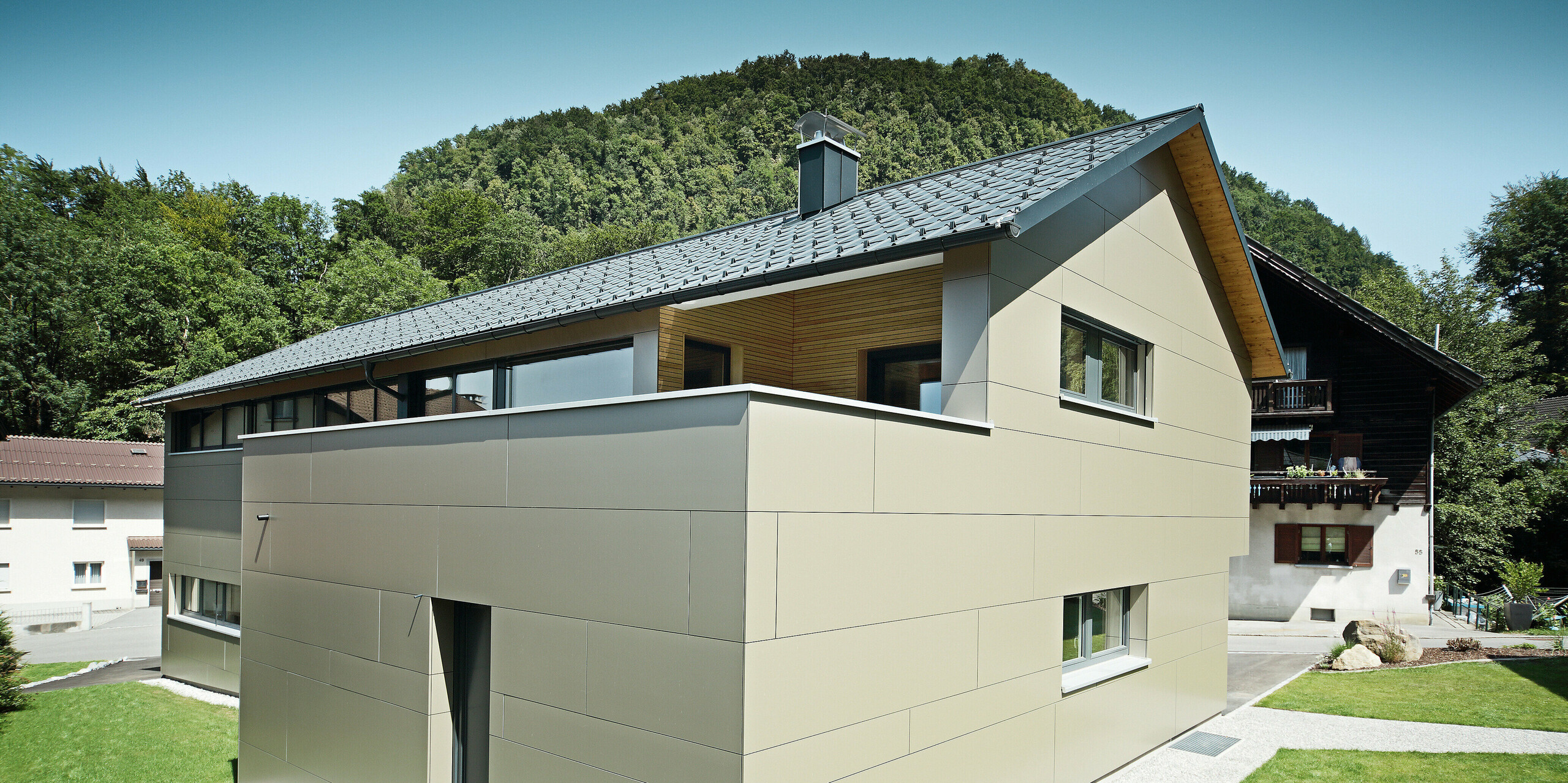 Einfamilienhaus in Hohenems mit einer stilvollen PREFA Fassade aus Aluminium Verbundplatten in Bronzemetallic und einem harmonierenden Dach aus PREFA Dachplatten in P.10 Anthrazit. Das Bild präsentiert das Gebäude in voller Pracht vor einem bewaldeten Hintergrund, wobei die saubere Linienführung und die Farbkombination der Materialien die zeitgenössische Architektur unterstreichen und für einen modernen, langlebigen und wartungsarmen Gebäudeschutz sorgen.