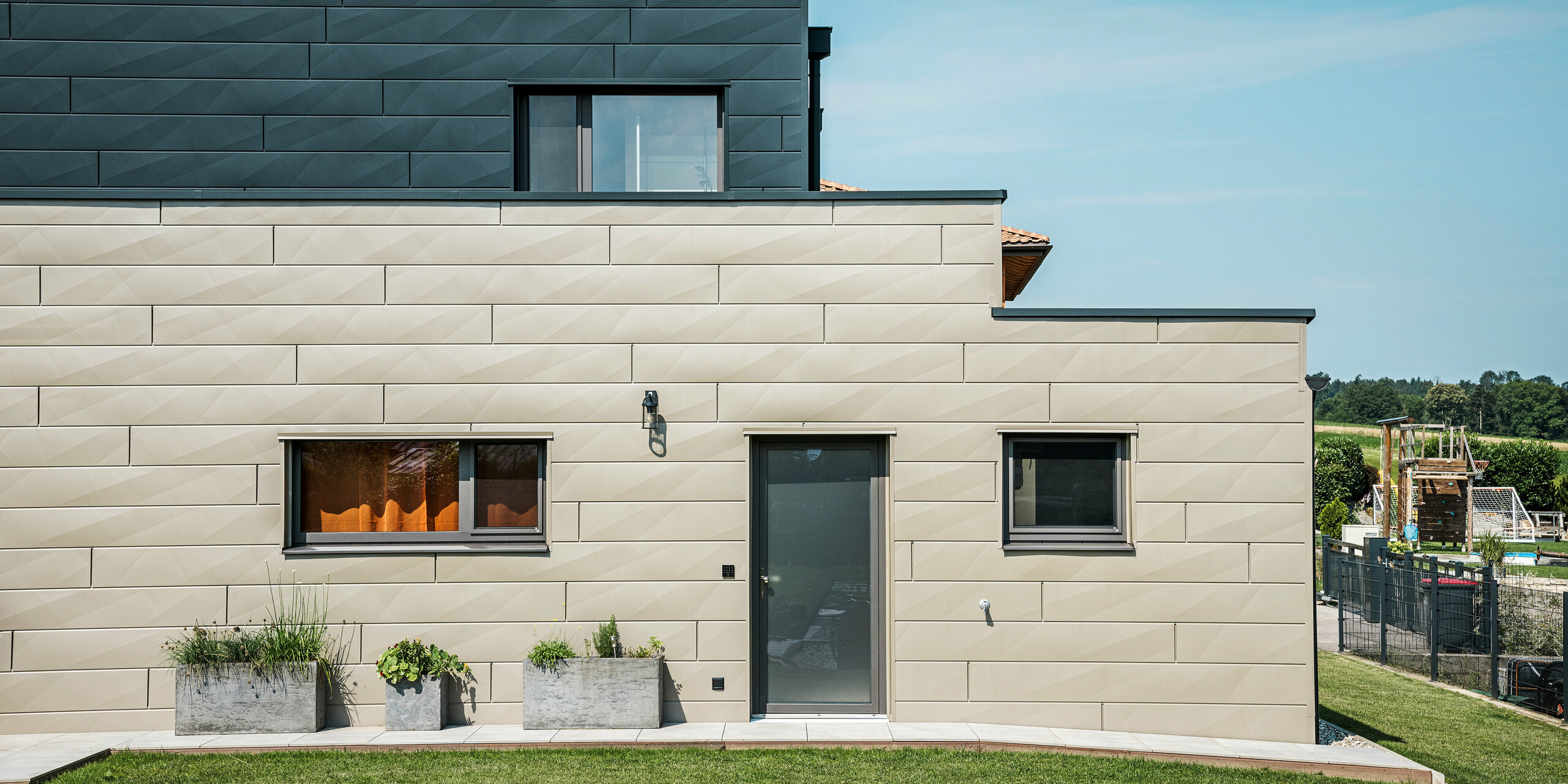 Vue latérale d'une maison familiale moderne à Wippenham, en Autriche, avec une façade en aluminium PREFA bicolore. La façade est conçue avec des panneaux de façade Siding.X dans les couleurs bronze et anthracite, qui assurent un look élégant et contemporain. Les lignes claires et les différentes couleurs soulignent le design moderne de la maison, tandis qu'un jardin bien entretenu complète l'ensemble harmonieux.