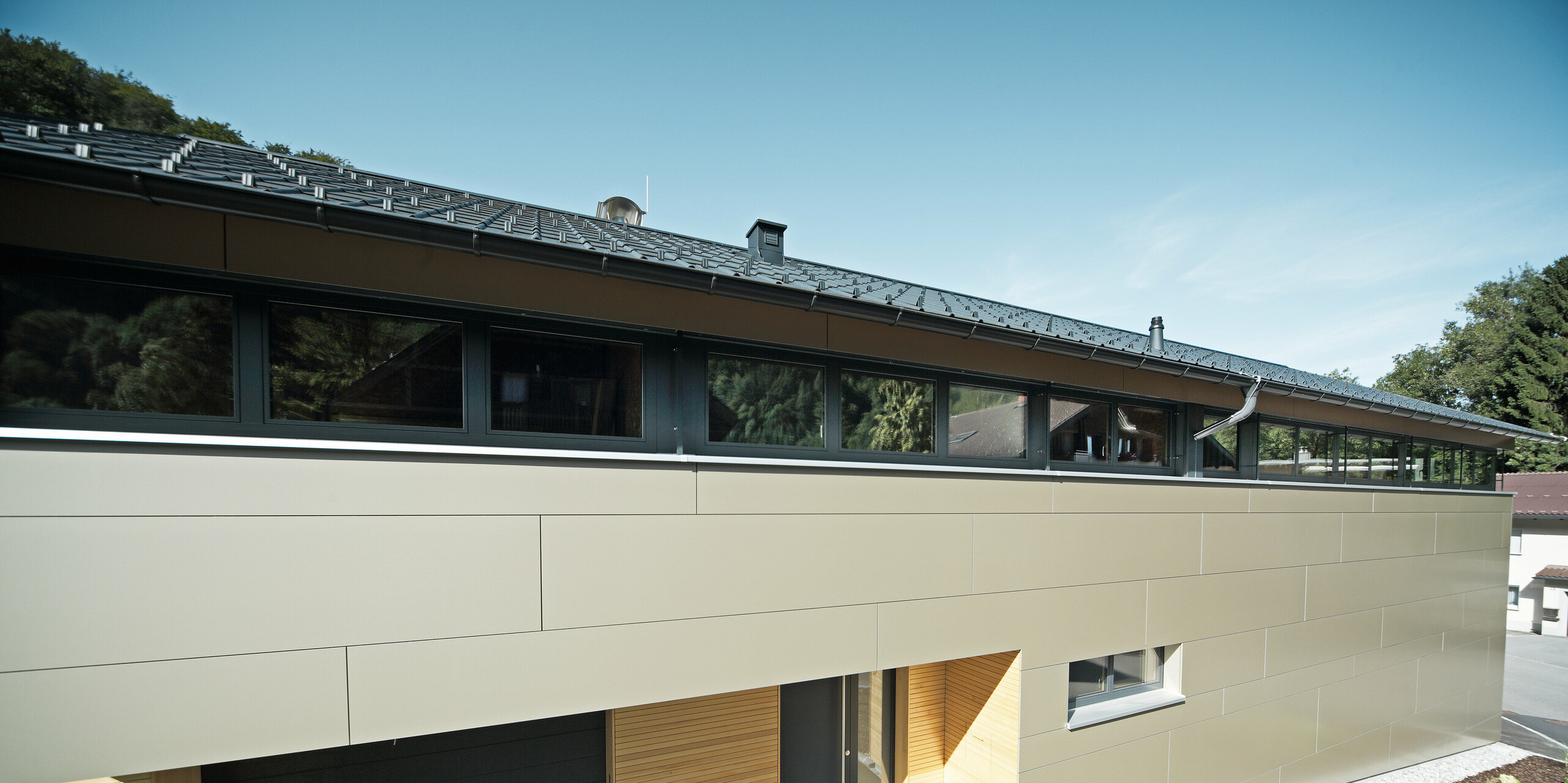 Frontansicht eines Einfamilienhauses in Hohenems, das mit PREFA Aluminium Verbundplatten in der Farbe Bronzemetallic verkleidet ist. Die horizontale Linienführung der Fassadenplatten schafft eine visuelle Breite und Struktur, während die Holzelemente im Eingangsbereich einen natürlichen Kontrast setzen. Das Dach ist mit PREFA Dachplatten in P.10 Anthrazit gedeckt, die perfekt mit der Fassade harmonieren und das moderne Design des Hauses unterstreichen.