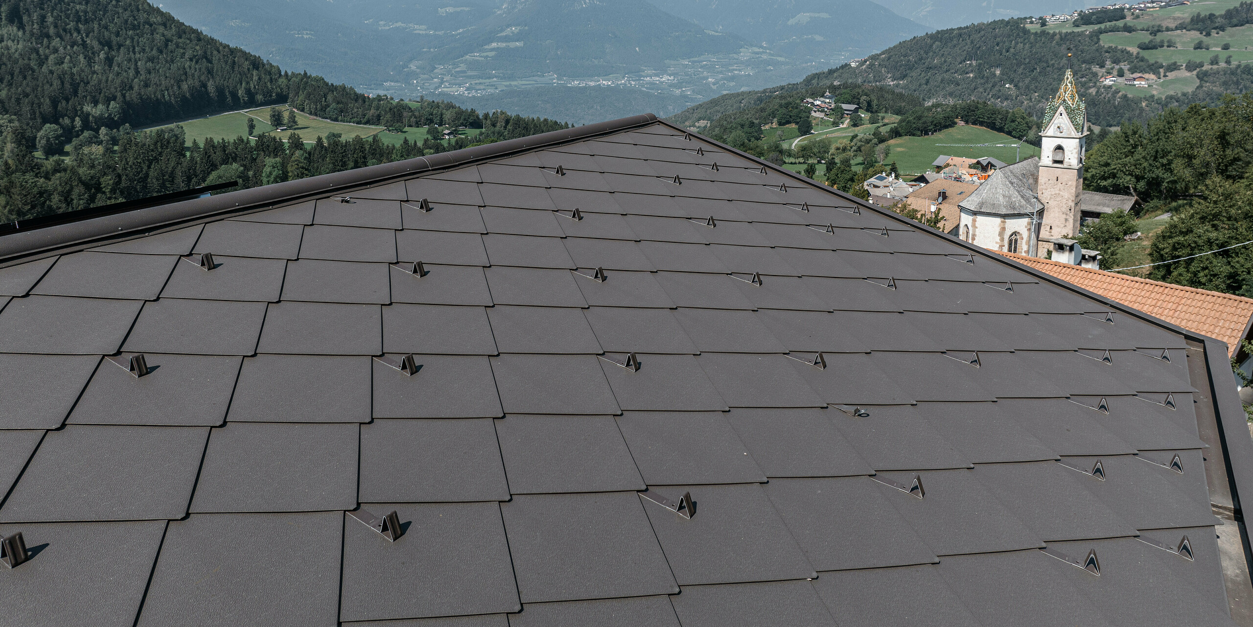 Detailní záběr střechy z hnědých hliníkových šablon PREFA v jihotyrolském Möltenu s působivým výhledem na okolní údolí a hory. Plechové kosočtverce nabízejí ochranu i estetiku a harmonicky zapadají do přírodní krajiny. V pozadí je vidět malebný vesnický kostel s charakteristickou věží, obklopený zelenými kopci a zemědělskou půdou. Pravidelně upevněné sněhové zábrany na střeše mají zabránit sesuvu střešních lavin. Systém ochrany proti sněhu je také v barvě P.10 tmavě hnědé a harmonicky zapadá do celkového obrazu díky jednotné barvě.