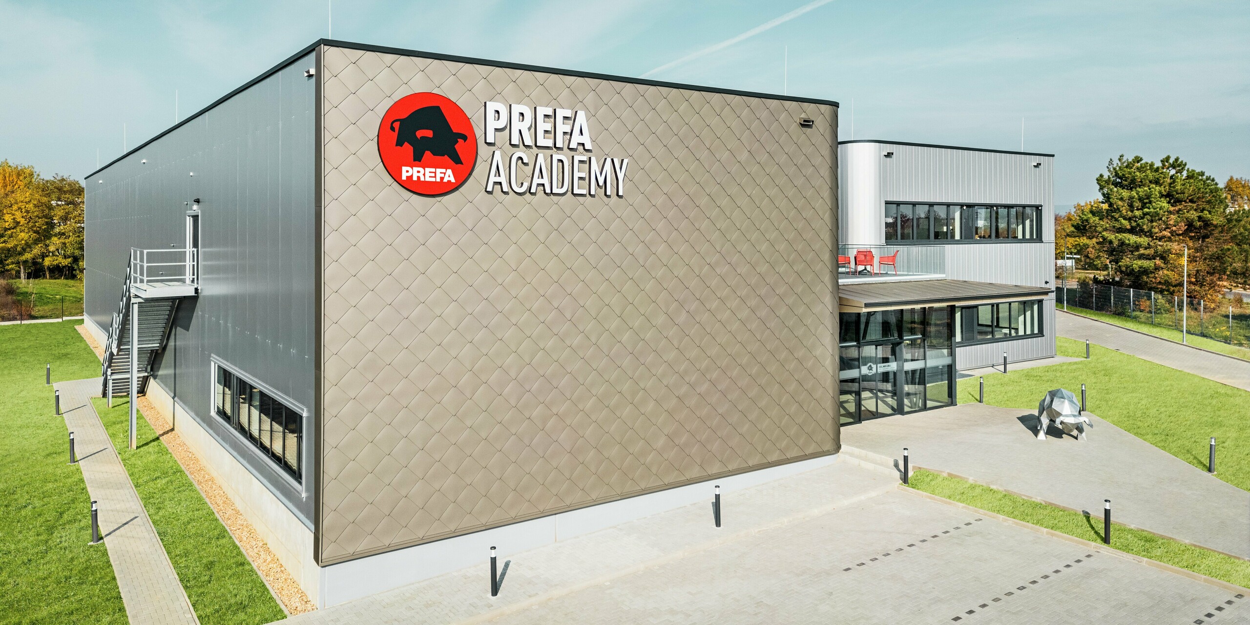 Die PREFA Academy in Törökbálint präsentiert sich mit einer markanten Fassade aus PREFA Wandrauten 44 × 44 in Bronze und PREFA Sidings in Silbermetallic. Diese hochwertigen Aluminiumprodukte sorgen für eine moderne Optik, extreme Witterungsbeständigkeit und Langlebigkeit. Das große PREFA Logo an der Fassade zum Parkplatz und die Stierskulptur am Weg zum Eingang unterstreichen die Stärke der Marke.